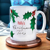 A personalised Christmas mug with the message "Holly's Christmas Tea" and illustrations of holly leaves printed onto it.
